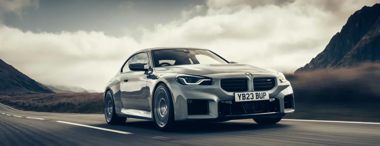 A silver BMW M2 performance coupe parked on an open road under a dramatic, cloudy sky.